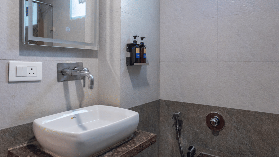 Modern hotel bathroom with a wall-mounted mirror, white basin, and subtle grey tile work under bright lighting at Pravasa Stay, Vadodara.