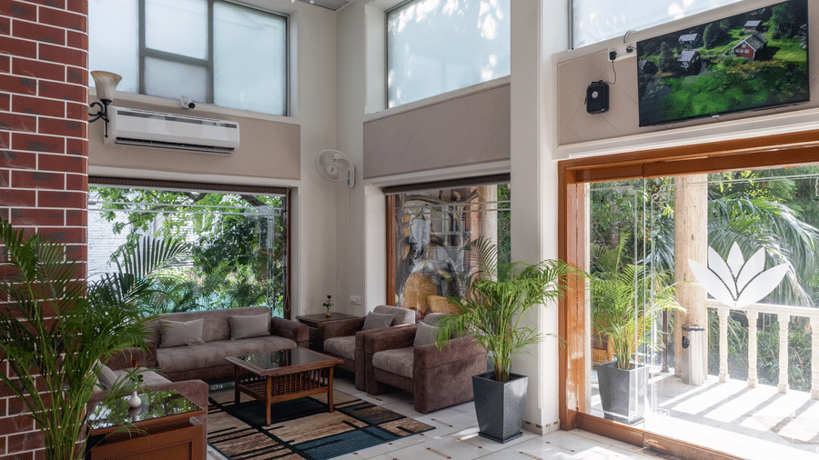 Bright, sunlit interior space with white tiled floors, high windows, and indoor plants, leading to an outdoor area at Pravasa Stay, Vadodara.