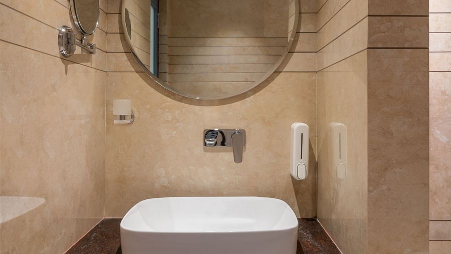 A bathroom features a round wall-mounted mirror above a wash basin | Hotel Ganga Maiya