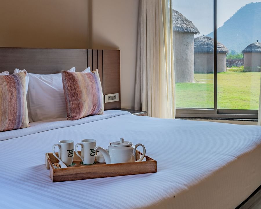 An image of a double size bed where a tea tray is placed and next to the bed there is a huge window from where outdoor scenery can be enjoyed  - Ananta Spa and Resort, Ajabgarh