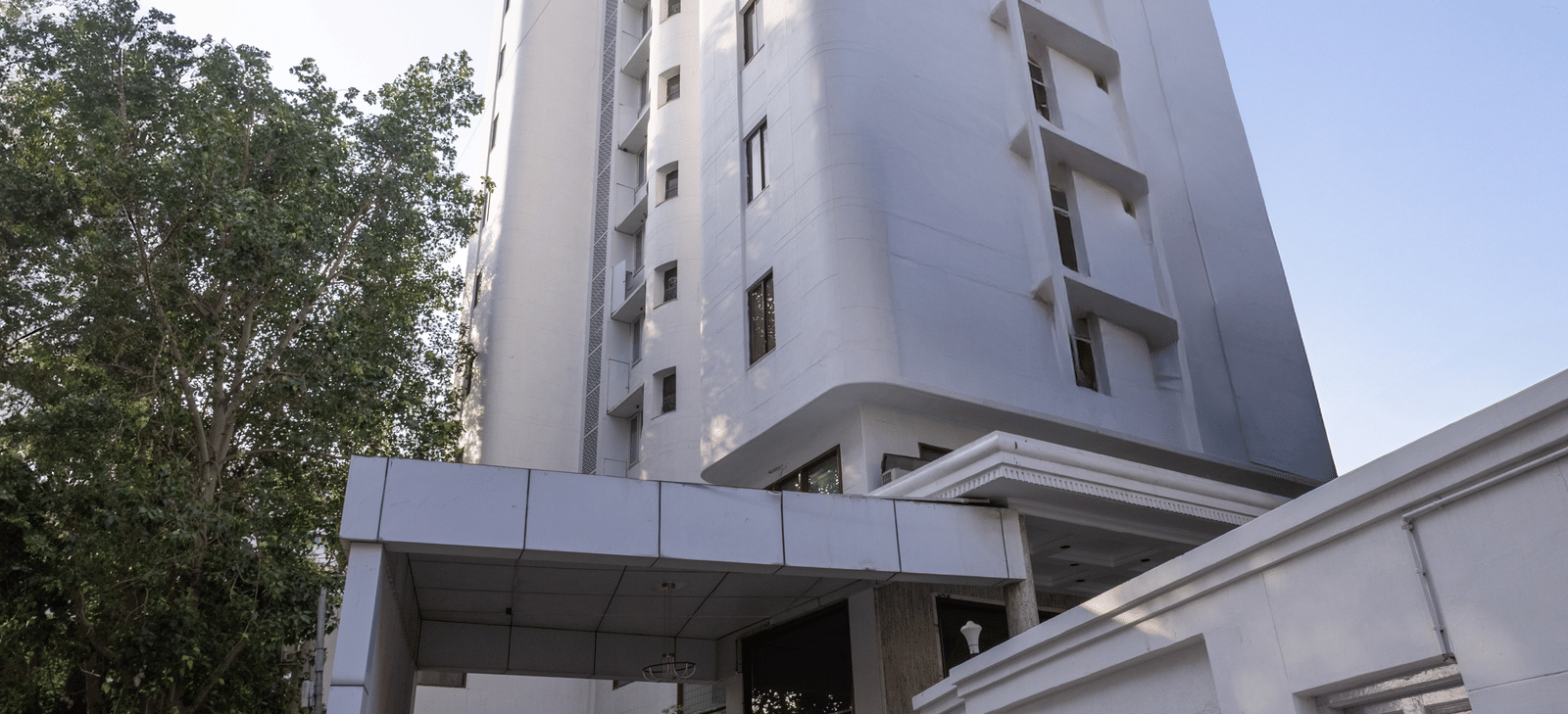 Exterior view of the tall, white hotel building standing against a clear sky, with a driveway leading to the entrance at Pravasa Stay, Vadodara.