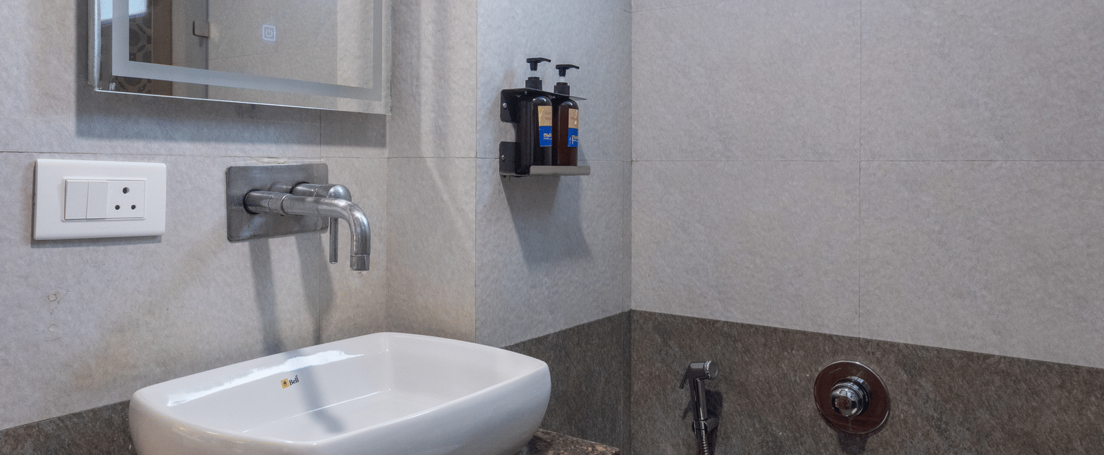 Modern hotel bathroom with a wall-mounted mirror, white basin, and subtle grey tile work under bright lighting at Pravasa Stay, Vadodara.