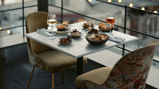 Two chairs and a table with food are positioned on a balcony overlooking a city.