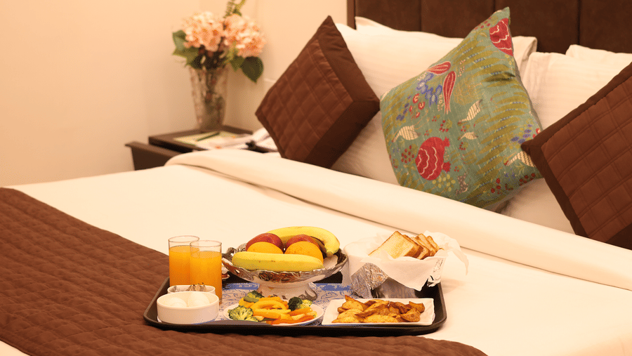 A clean, well-lit hotel guest room with a bed and a breakfast tray, with a nightstand and lamp visible.