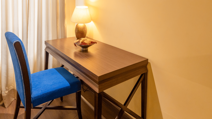 A desk with a chair, table, lamp, and wall art placed against a wall inside a room at Indraprastha Resort & Spa, McLeod Ganj.