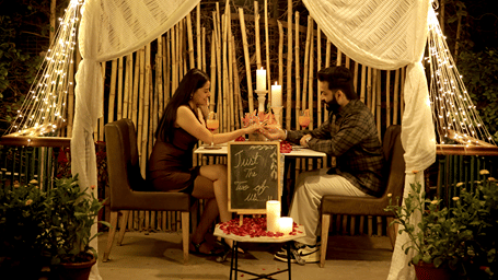 A couple dining at an intimate outdoor table beneath a draped canopy, surrounded by candles, string lights, bamboo decor, and plants.