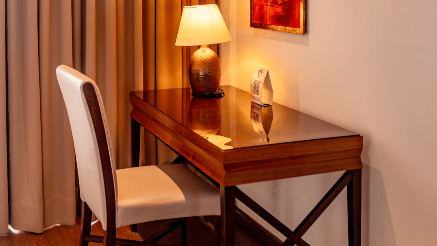 A desk with a chair, table, lamp, and wall art placed near curtains inside a room at Indraprastha Resort & Spa, McLeod Ganj.