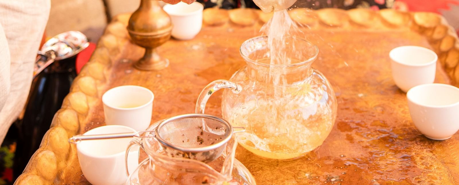 Outdoor tea setup with a person pouring tea into a teapot, while a lady sits behind observing the tea making attentively.