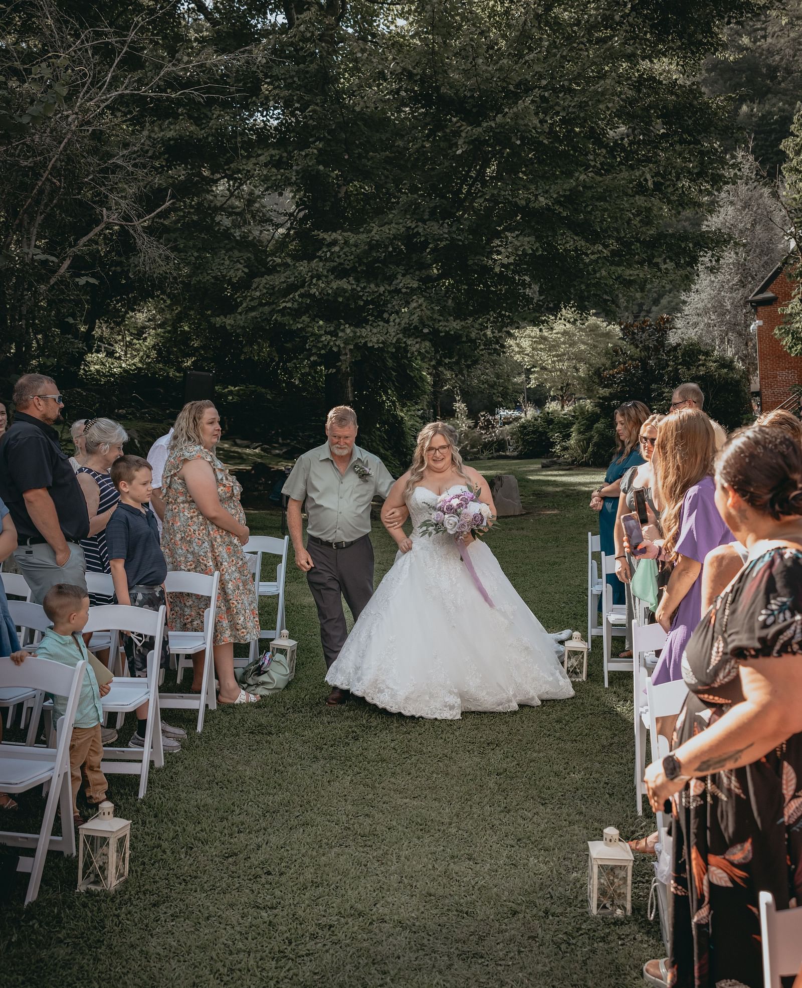 Bride walking down the aisle holding a bouquet and her dad at the Historic Tapoco Lodge. On either side of the aisle there are people people standing for the bride next to their chairs. 