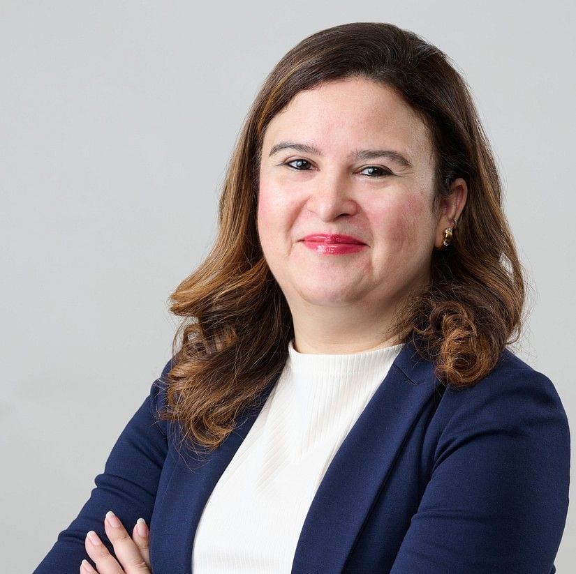 Mielle Batliwala with brown hair, wearing a navy blazer and white shirt, standing with arms crossed, smiling slightly.