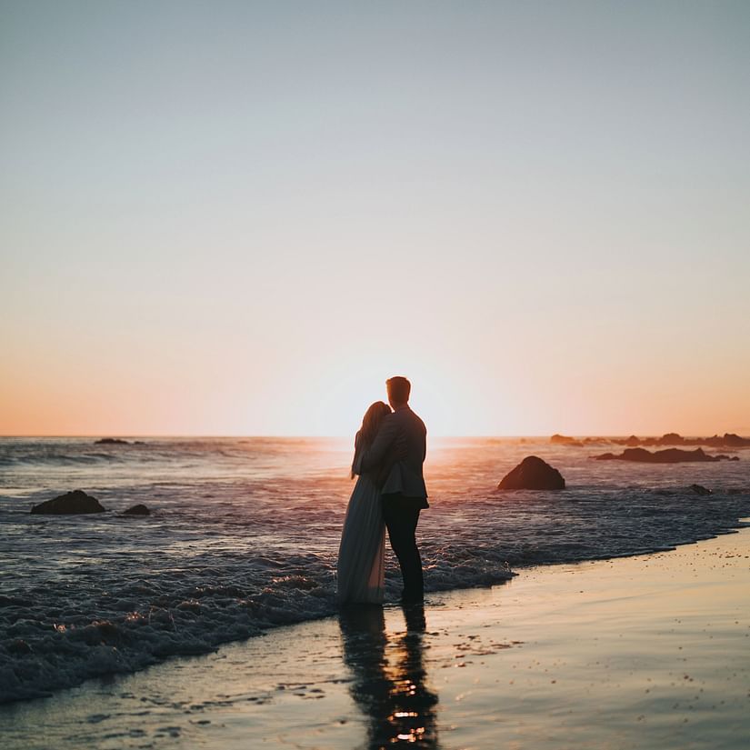 a couple hugging while standing and looking at the sun set on a beach while overlapping waves hits their feet