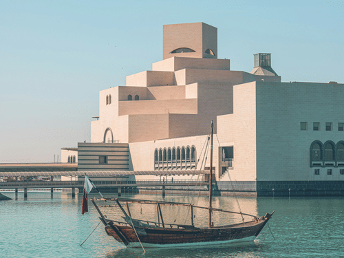 A traditional wooden boat, or dhow, floats on turquoise water in front of the Museum of Islamic Art in Doha, Qatar. A must visit in your 2 Days in Doha.