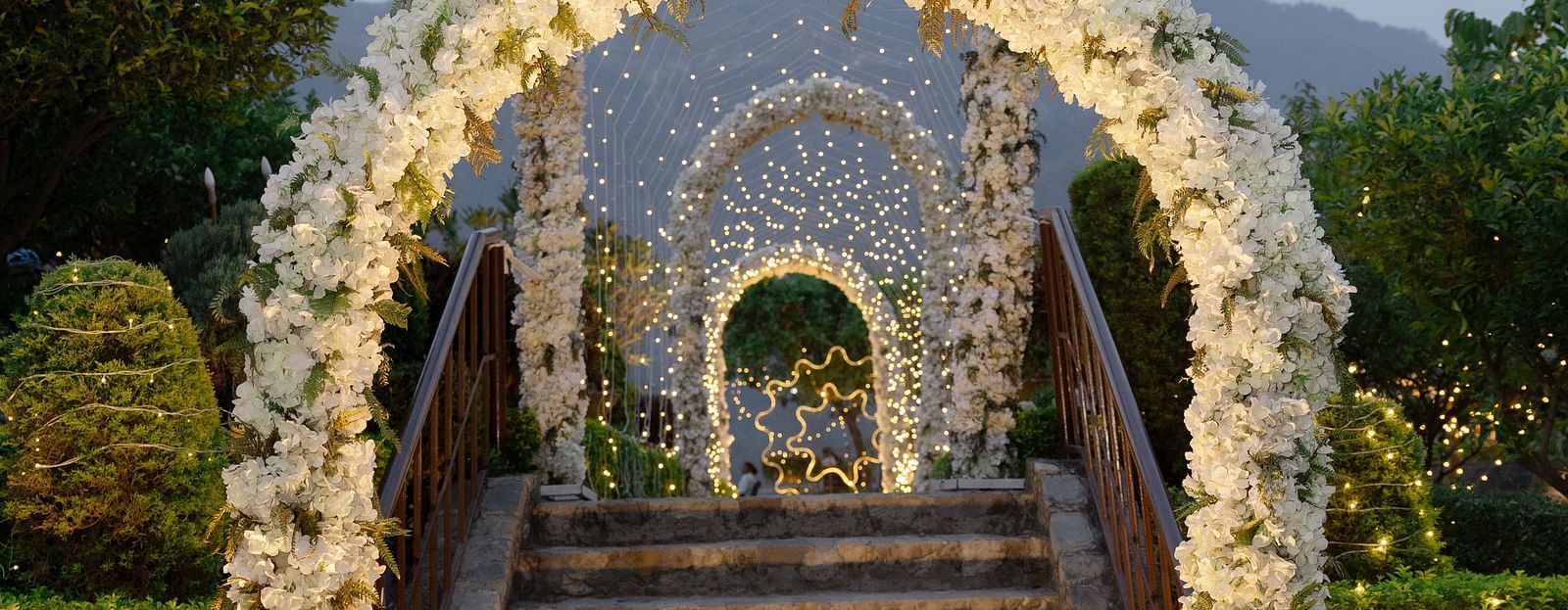 A grand floral archway leading to stairs, adorned with flowers and illuminated by soft lighting, 
