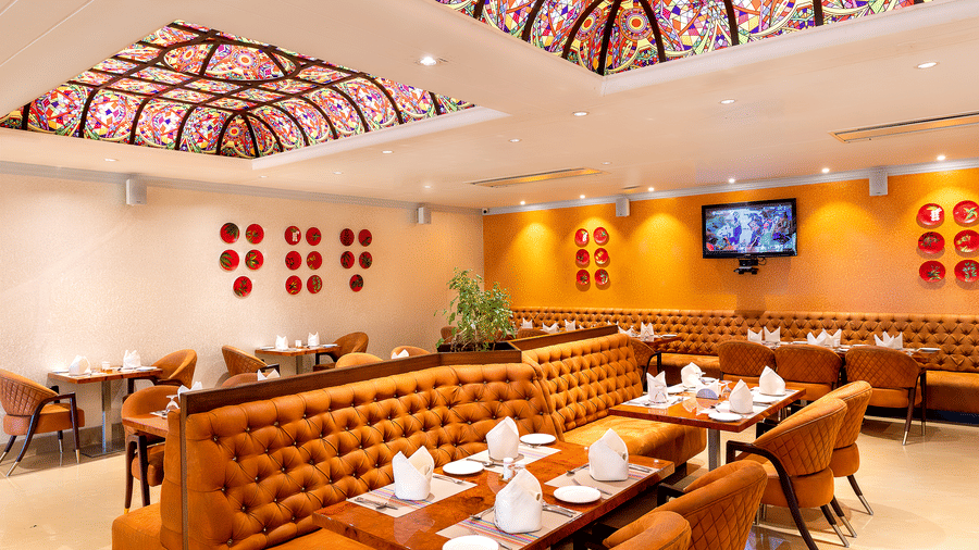 Interior view of the brightly lit restaurant showing the corner seating, orange booths, and large decorative glass ceiling at Hotel Hukam's Lalit Mahal.