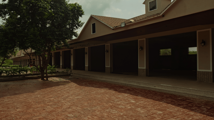 A wide brick-paved courtyard leads toward a long building with a covered walkway under a cloudy sky with green trees to the left at Jivana Wellness Retreat, Bhopal.