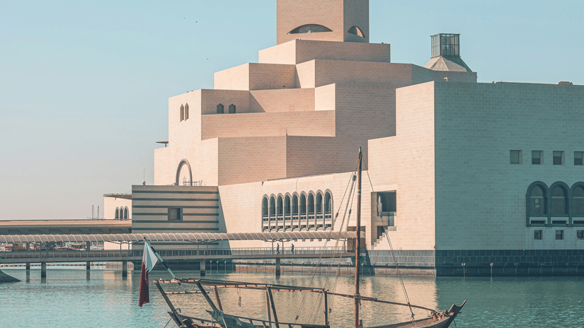 A traditional wooden boat, or dhow, floats on turquoise water in front of the Museum of Islamic Art in Doha, Qatar.