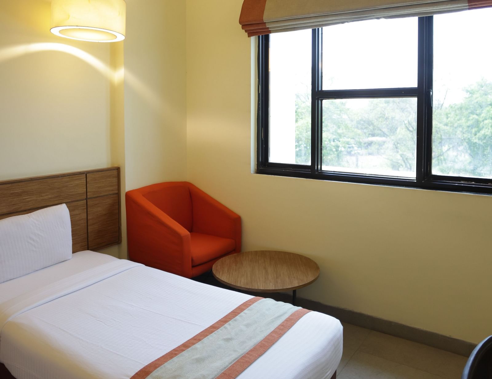 a close up shot of a comfortable single bed with a red cushioned armchair in the corner of the room with a coffee table in front of it  - Max Hotel Jabalpur