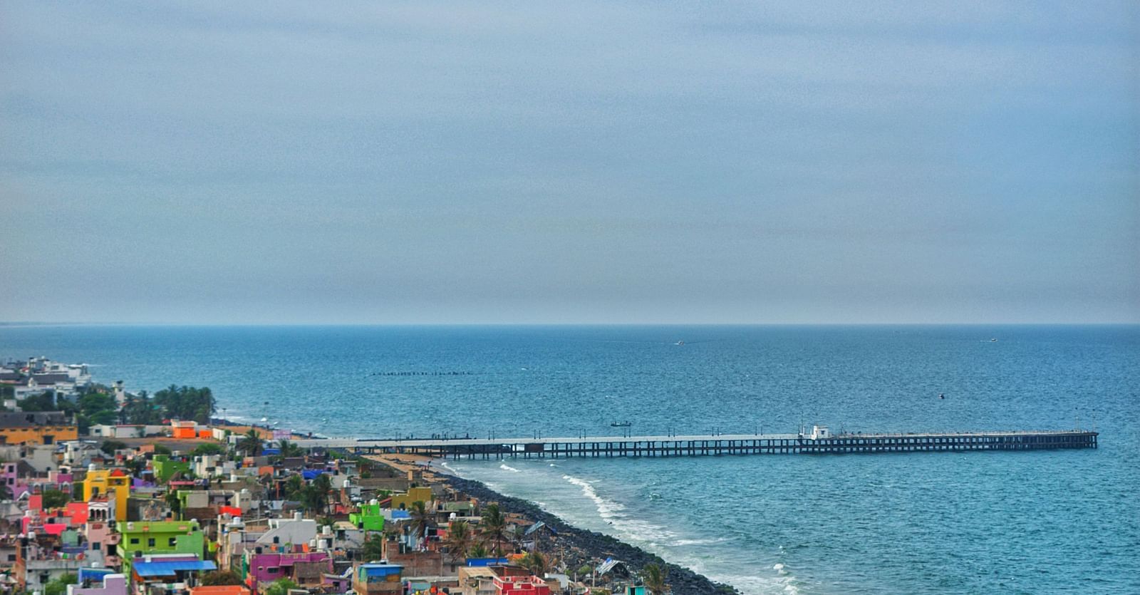 An aerial view of Pondicherry featuring colourful buildings clustered along the coastline next to the Bay of Bengal and a long pier extending into the sea.