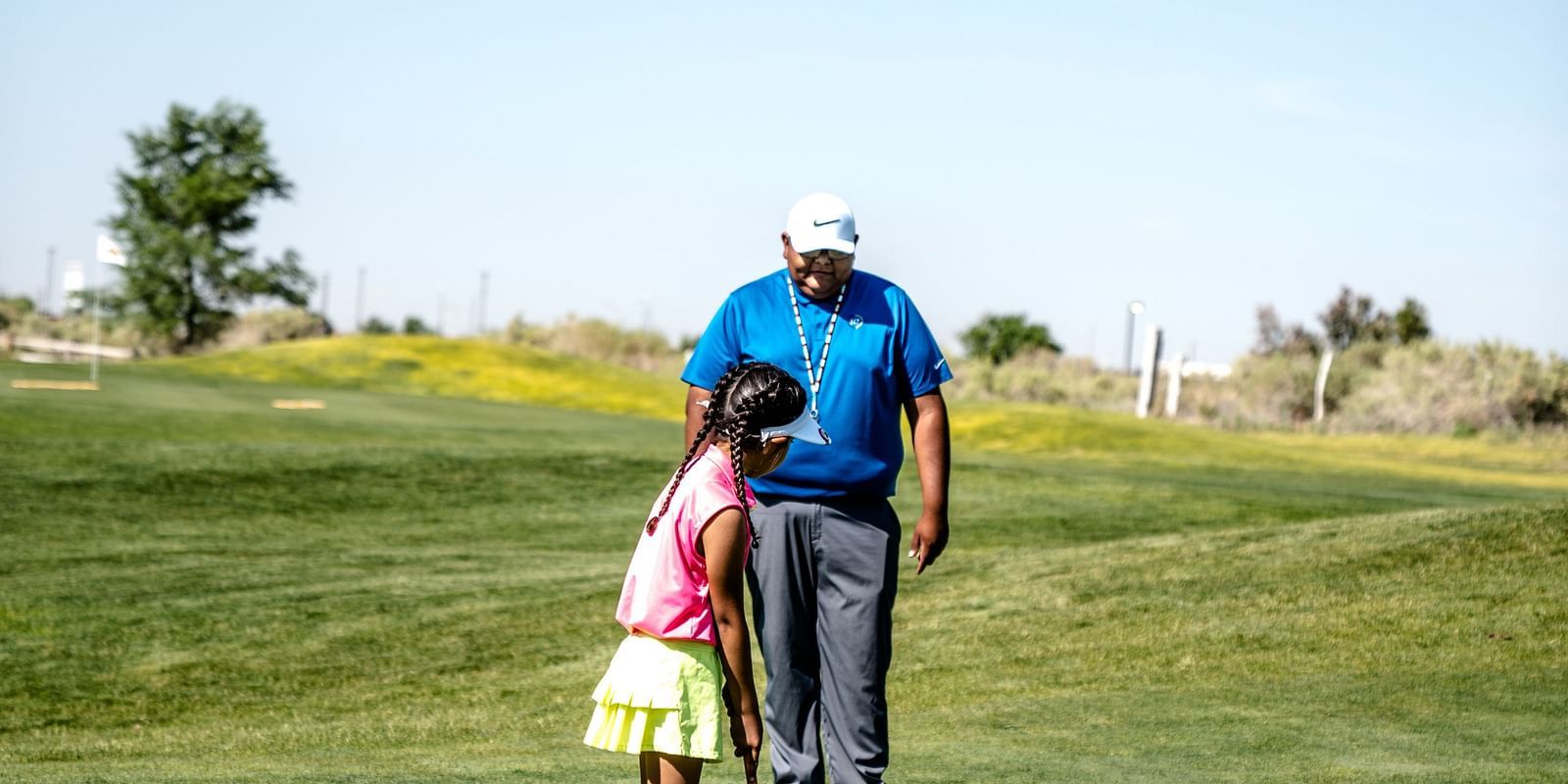 A young girl practicing her putting on a golf course, with an adult coach guiding her in the background.