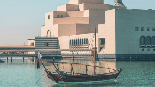 A traditional wooden boat, or dhow, floats on turquoise water in front of the Museum of Islamic Art in Doha, Qatar. A must visit in your 2 Days in Doha.