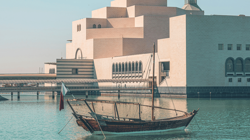 A traditional wooden boat, or dhow, floats on turquoise water in front of the Museum of Islamic Art in Doha, Qatar.