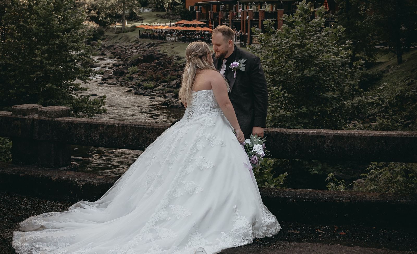 Just married couple looking at each other, meaning to pose for the camera for their wedding shoot. At the background there is the Historic Tapoco Lodge surrounded by tall trees, hills,  and a stream running by in front of the property.