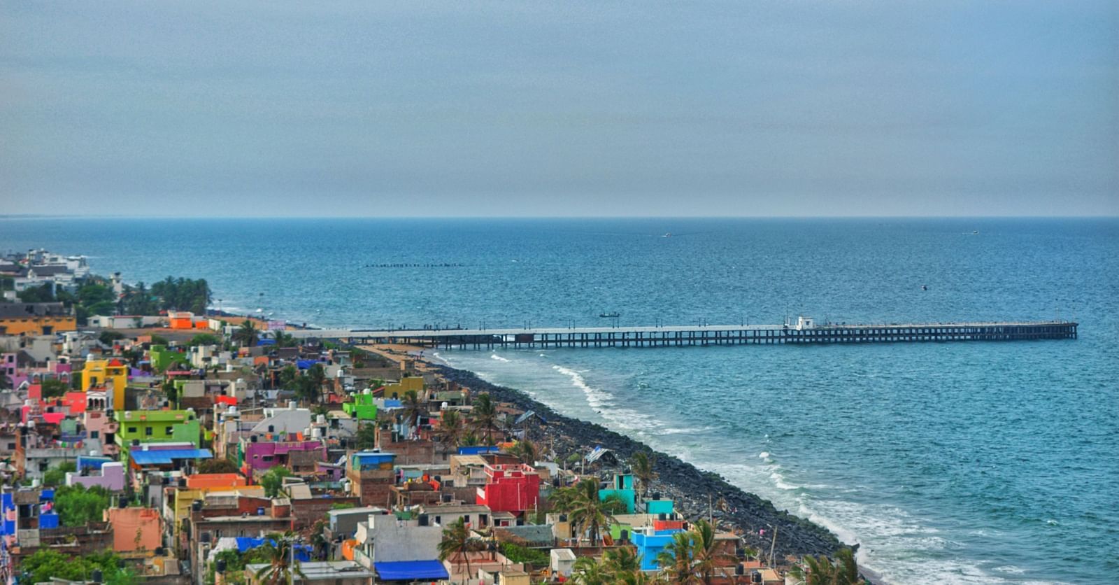 An aerial view of Pondicherry featuring colourful buildings clustered along the coastline next to the Bay of Bengal and a long pier extending into the sea.
