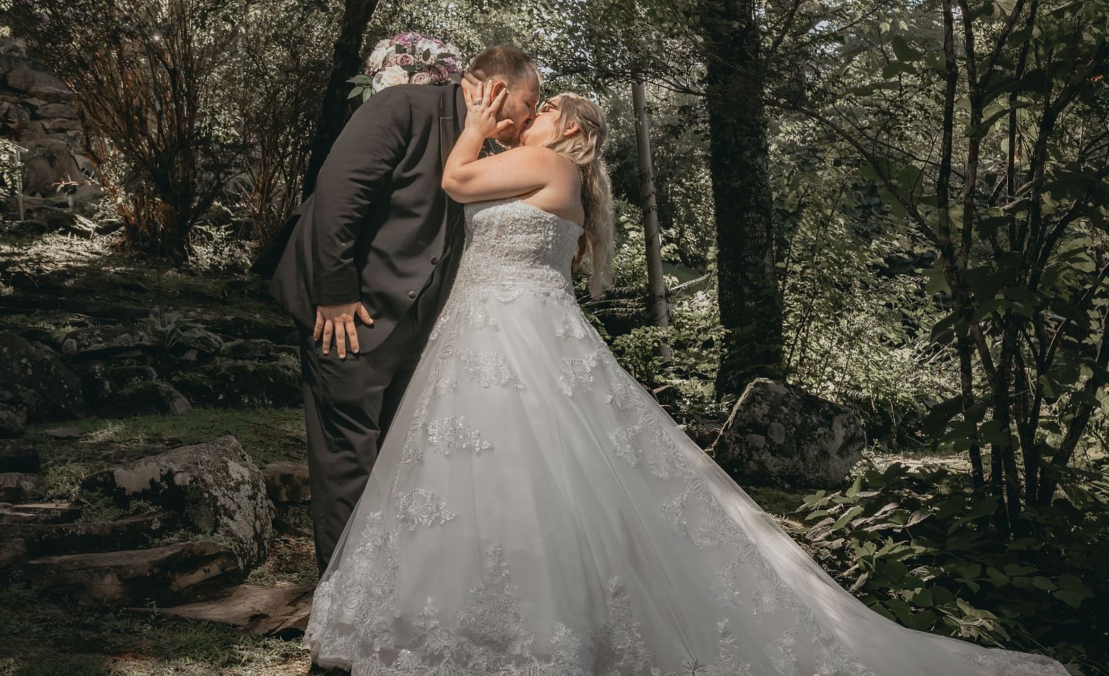A couple in wedding attire kissing each other in a forest covered with tall trees from where sunlight is peaking through.