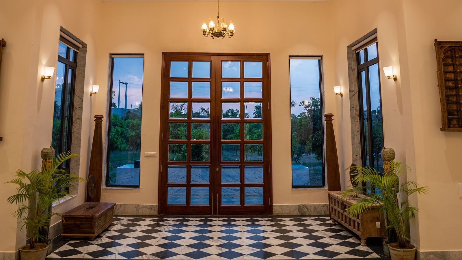An image of a lobby decorated with plants on the corner of the room, a pot filled with flowers placed on a table and there is a chandelier light on top - Rajbagh Wellness