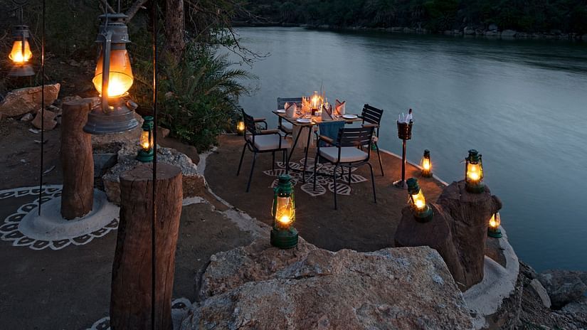 a table for four set up next to a lake and trees with lamps lighting the table - Chunda Shikar Oudi