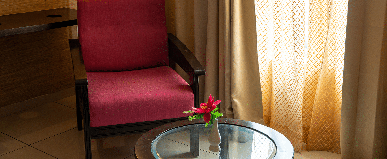 A small seating area with a glass-top table and a armchair next to a window with golden curtains - Hotel Udai Median, Udaipur.