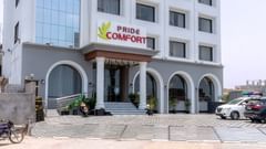 A facade of Pride Elite Dwarka featuring multi storey building and at the entrance there is a logo of the hotel. The front yard has two wheeler and four wheeler parked.
