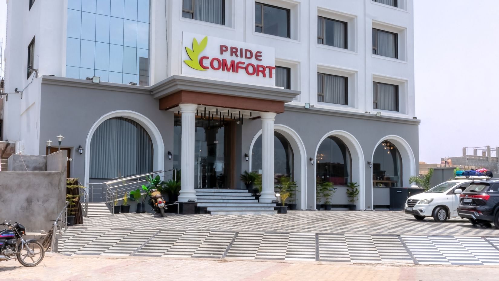 A facade of Pride Comfort Dwarka featuring multi storey building and at the entrance there is a logo of the hotel. The front yard has two wheeler and four wheeler parked.