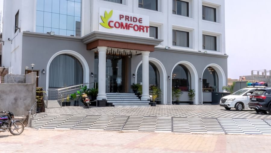 A facade of Pride Comfort Dwarka featuring multi storey building and at the entrance there is a logo of the hotel. The front yard has two wheeler and four wheeler parked.