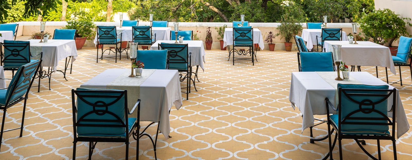 Another view of an outdoor dining area at Royal Heritage Haveli with tables and chairs visible through tree foliage.
