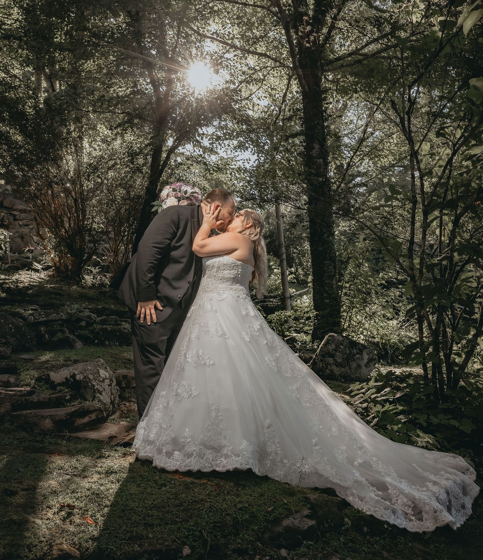 A couple in wedding attire kissing each other in a forest covered with tall trees from where sunlight is peaking through.