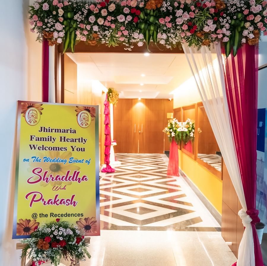A decorated entrance to a wedding venue with floral arrangements, drapes, and a welcome sign. The sign reads Mangalya Mandapam and the name of the bride and groom.