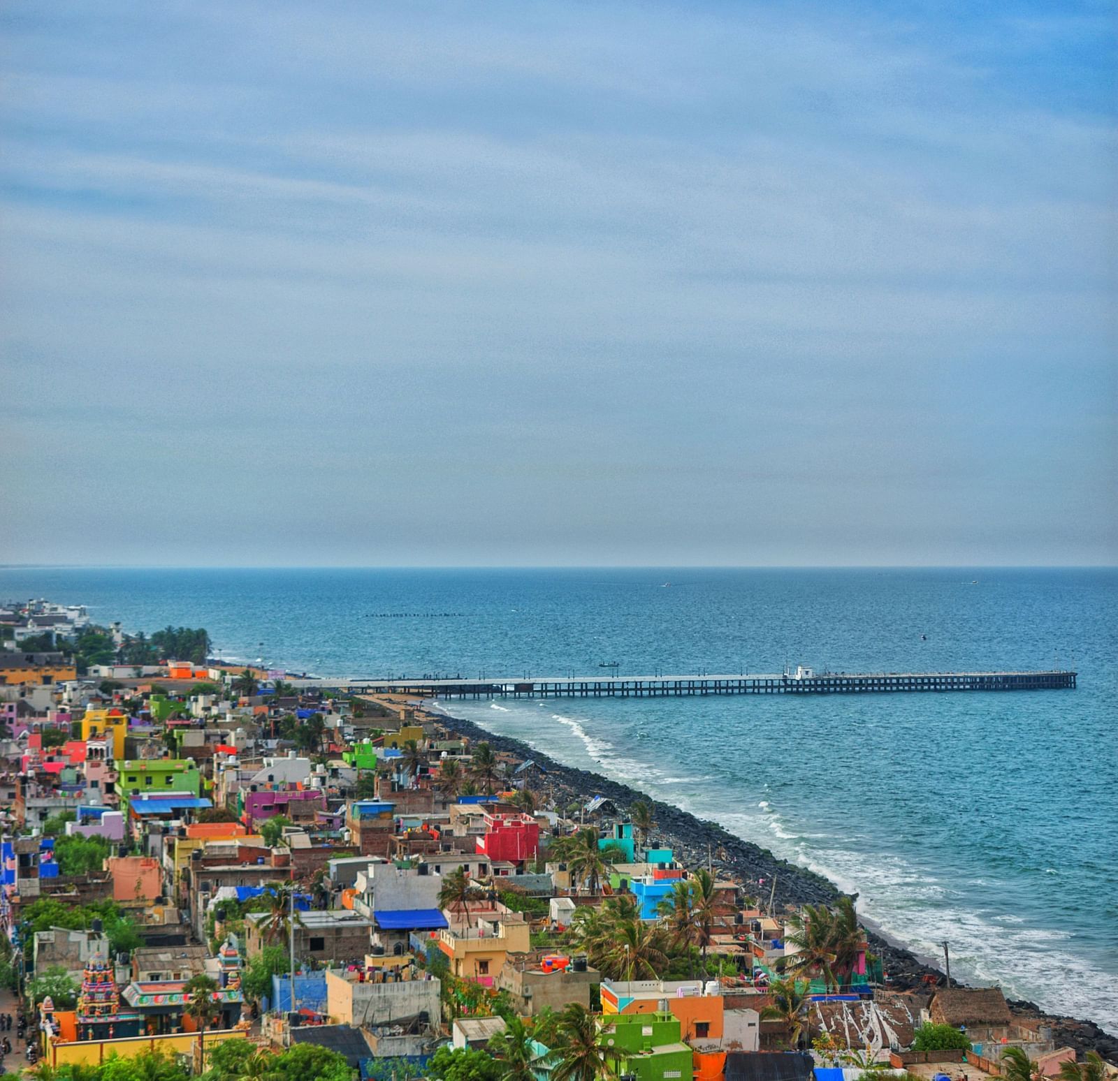 An aerial view of Pondicherry featuring colourful buildings clustered along the coastline next to the Bay of Bengal and a long pier extending into the sea.