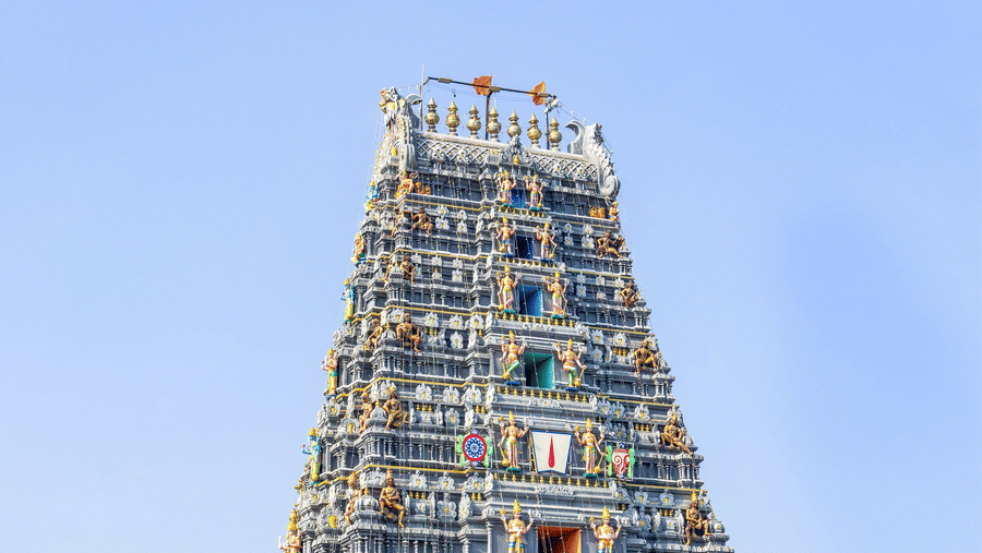 An ornate, tall South Indian temple tower stands prominently under a clear blue sky.