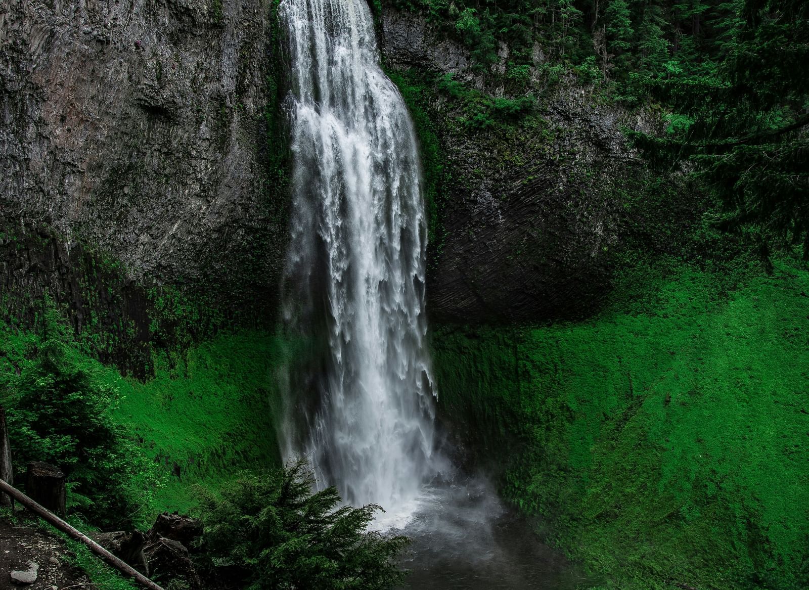 A tall, narrow waterfall plunges down a rocky cliff, surrounded by dense green forest.