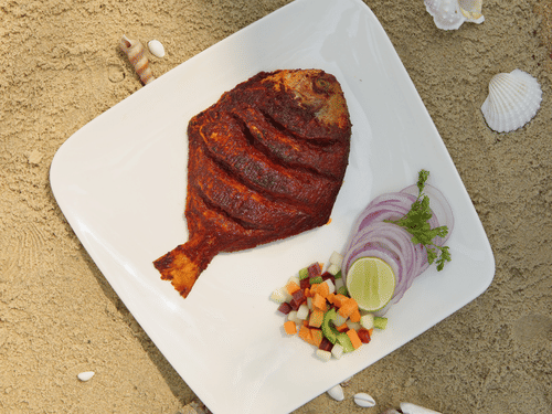 A top-down view of a cooked, seasoned whole fish on a square white plate, accompanied by sliced onions and lime, resting on sand.