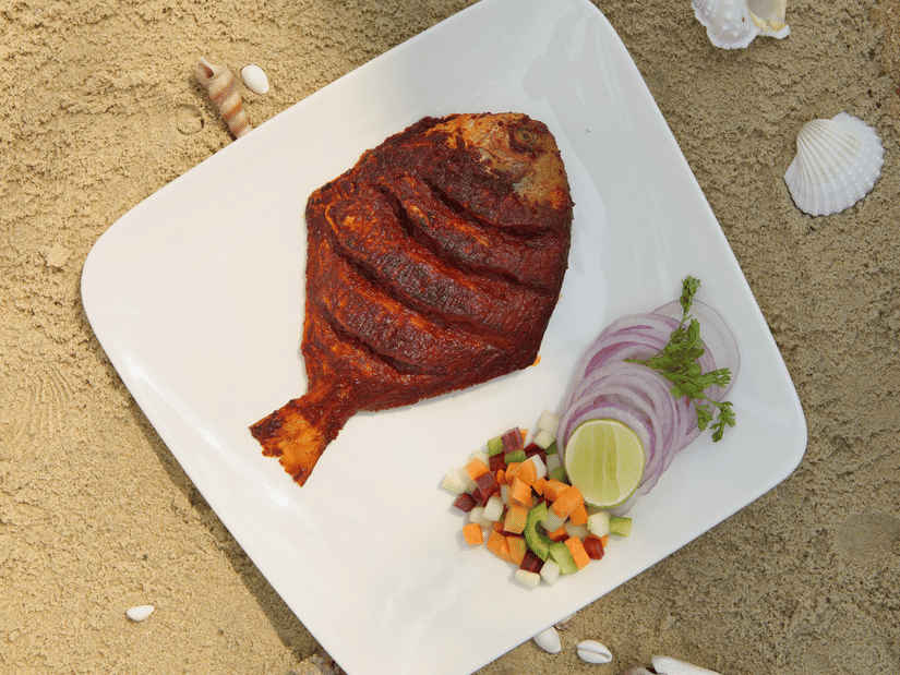 A top-down view of a cooked, seasoned whole fish on a square white plate, accompanied by sliced onions and lime, resting on sand.