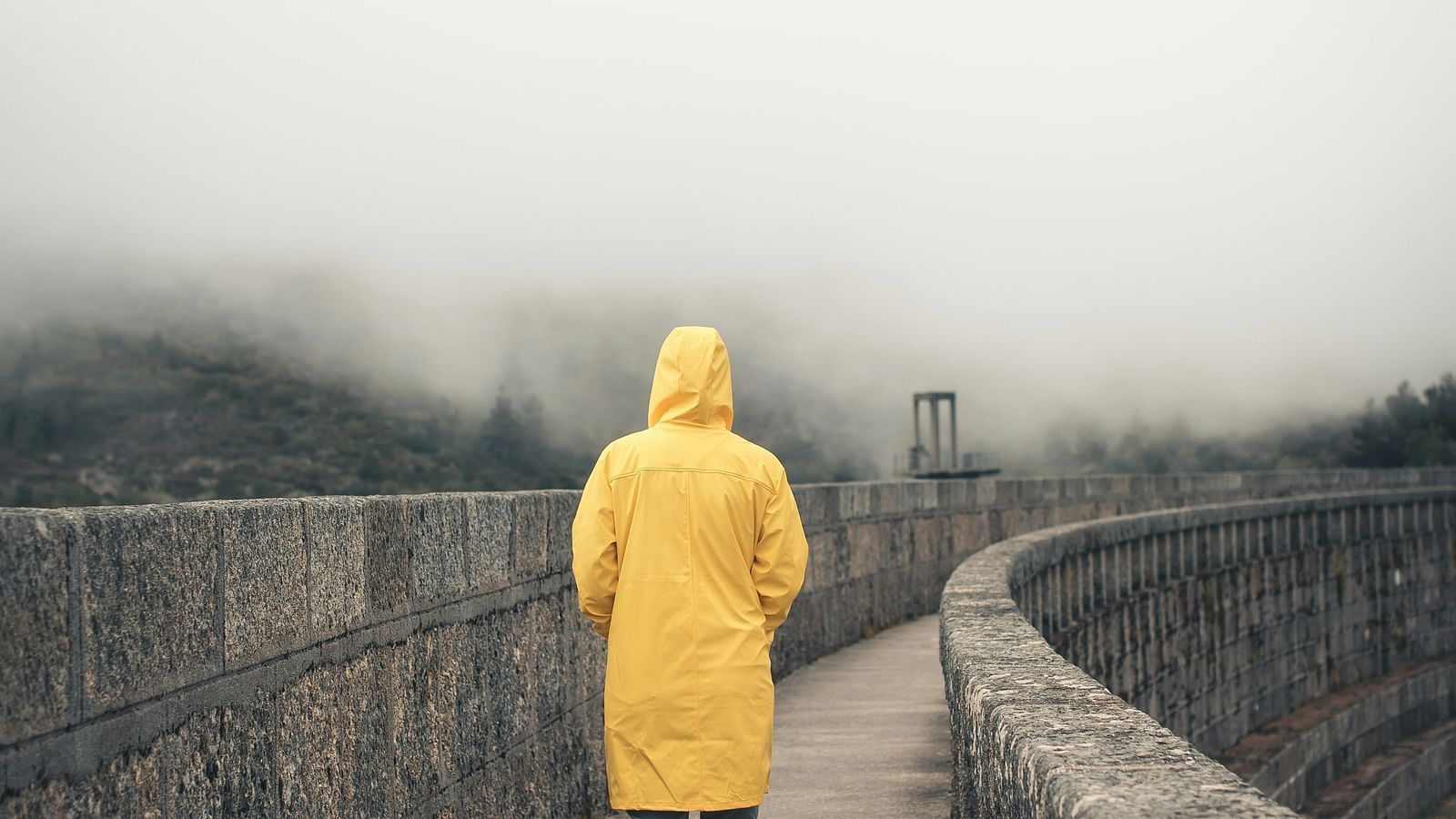An overview of a person wearing a yellow raincoat and walking on a pathway next to a dam.