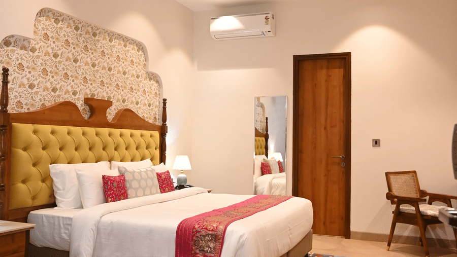 A guest room at Kadamb Kuteer with king bed, white linens, patterned pillows, a small seating area, and neutral-colored tiled flooring.