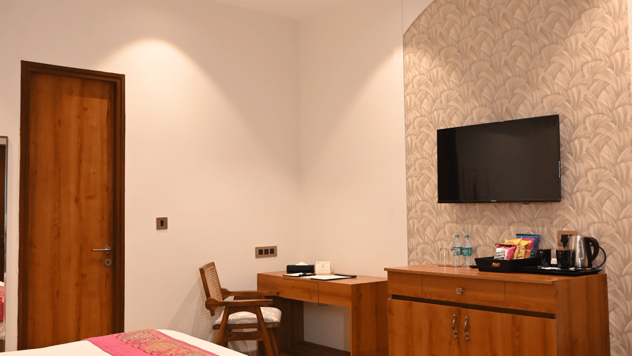 A guest room corner at Kadamb Kuteer showing a wooden desk, a wall-mounted TV, and a glimpse of the bed area with a patterned floral carpet.