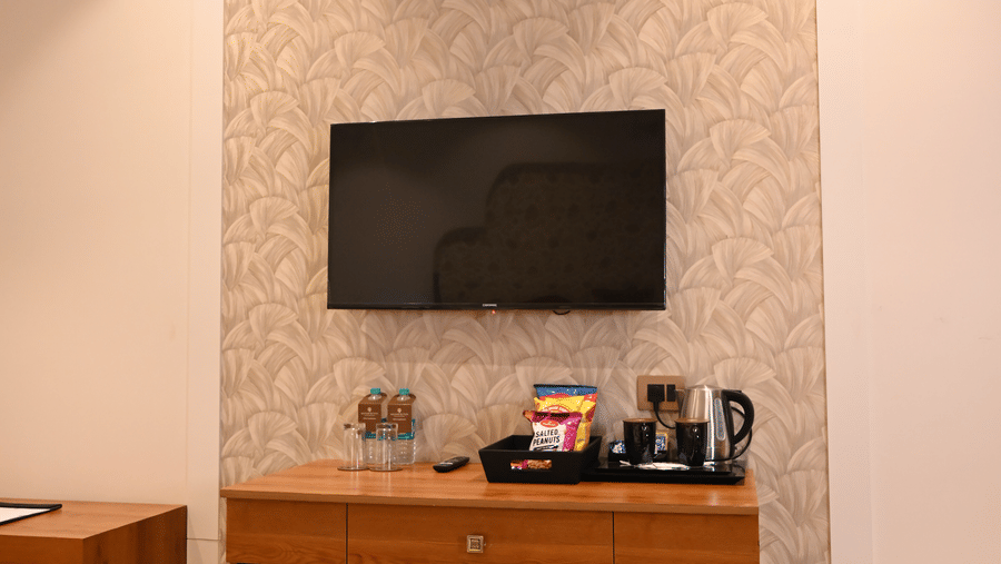A guest room interior at Kadamb Kuteer showing a wooden television console, a wall-mounted TV, and an arched recessed wall design.