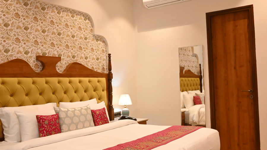 A view of a Kadamb Kuteer guest room featuring a double bed, a wooden wardrobe, and a seating area with a small table and patterned chairs.