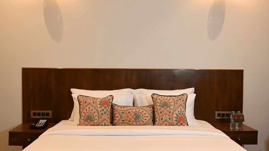 A wide view of a Kadamb Kuteer bedroom showing a king-sized bed with a dark wood headboard, white bedding, and wall-mounted reading lights.