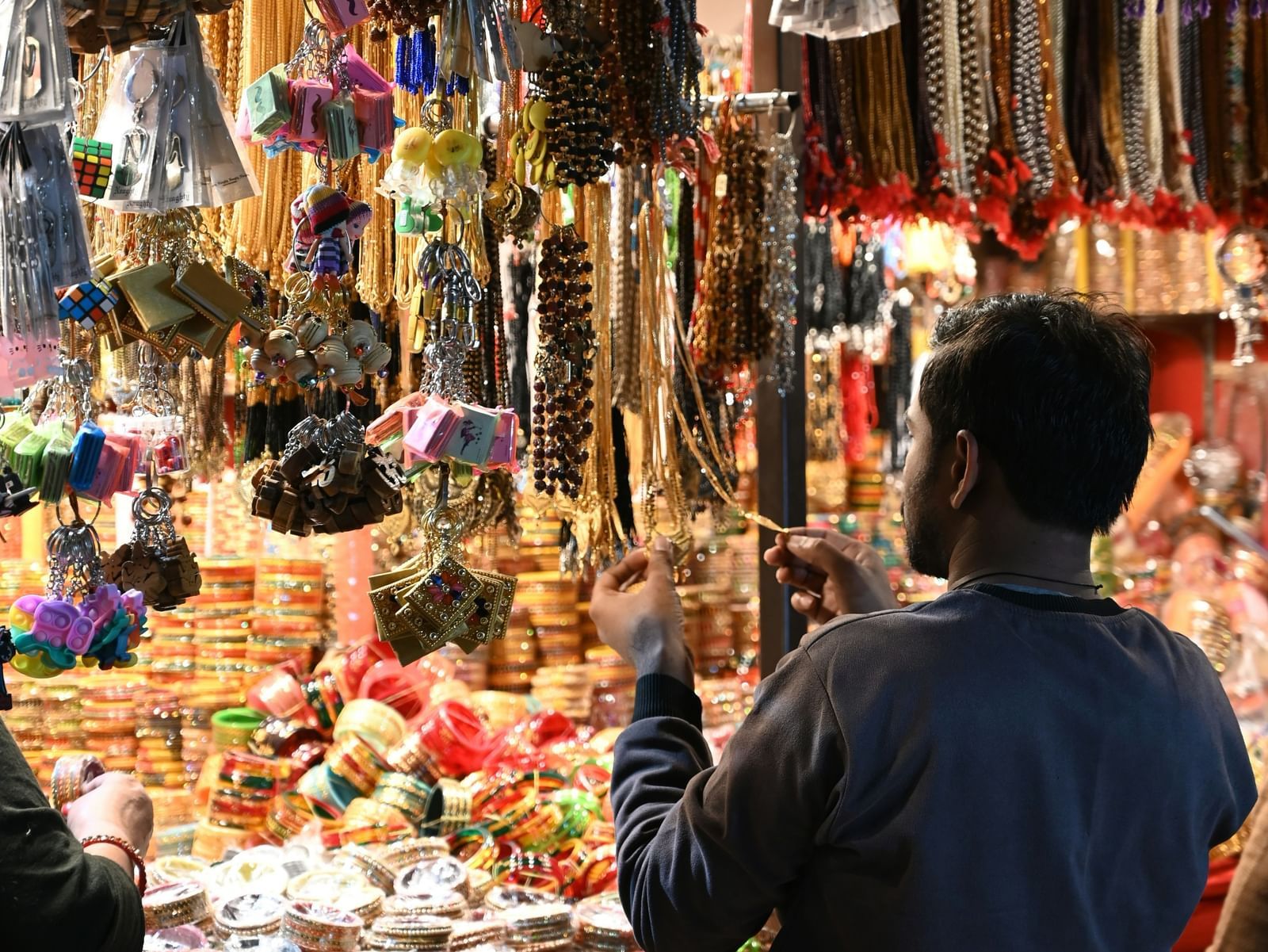 A man browses an overflowing market stall selling colourful stacks of bangles, beaded necklaces, and assorted keychains and trinkets hanging from the ceiling.