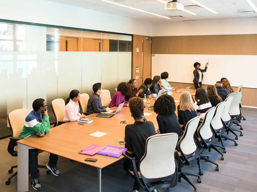 A diverse group of professionals seated around a conference table in a modern meeting room, listening to a presenter explaining points on a whiteboard during a team discussion.